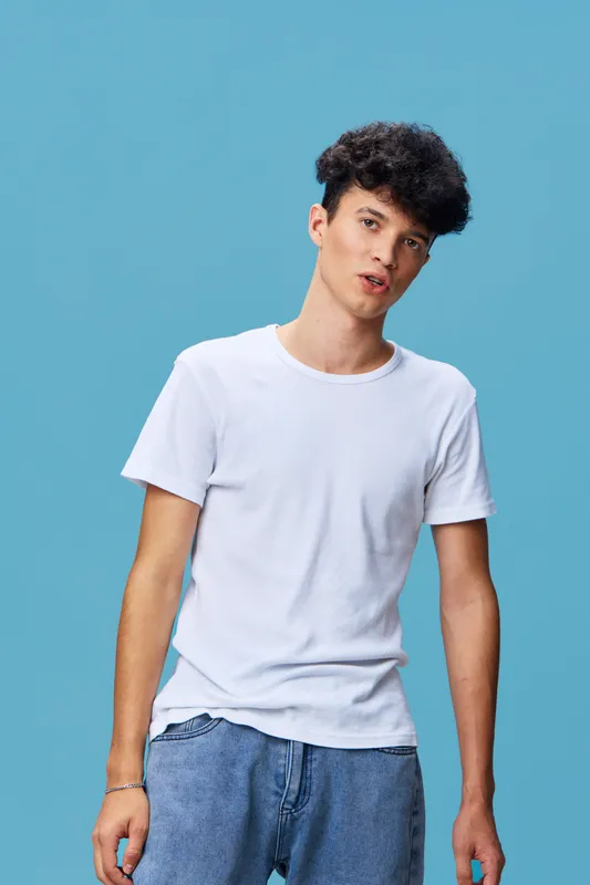 young man wearing casual white t-shirt and blue jeans standing against blue background with curly hair and relaxed expression