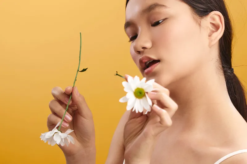 Asian woman delicately holds a white flower against a warm background, studying its petals with soft curiosity, creating a serene and artistic portrait of gentle beauty that captures light, texture,