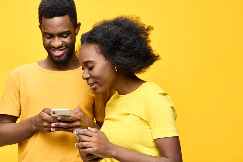 Young African couple enjoying their time together, smiling and interacting while using a smartphone, bright yellow background, summer vibes, joyful relationship