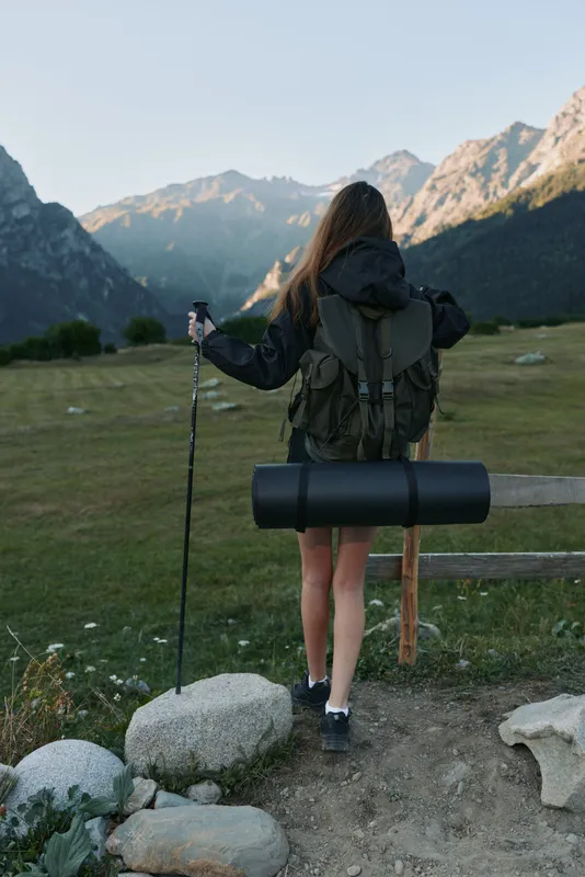 A lone hiker stands at a trailhead in a rugged alpine valley. She wears a backpack and uses trekking poles, ready to explore the scenic landscape.