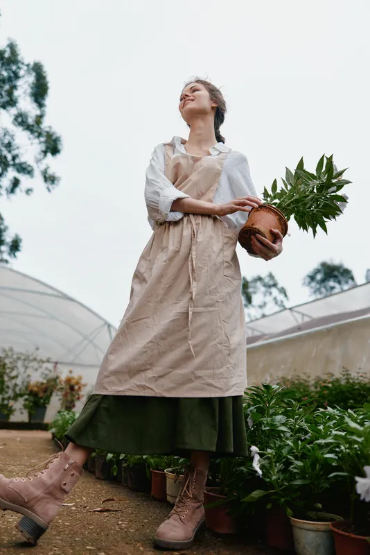 Woman in an apron holding a potted plant, showcasing a joyful expression in a lush greenhouse setting, reflecting the essence of gardening and nature