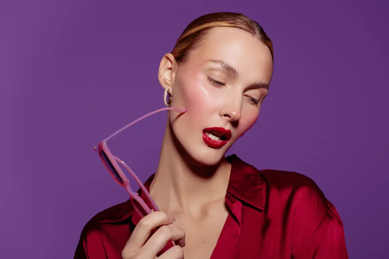 Portrait of a stylish woman with slicked hair and red lipstick holding pink glasses against purple background. Concept of fashion, beauty, elegance, and modern style.