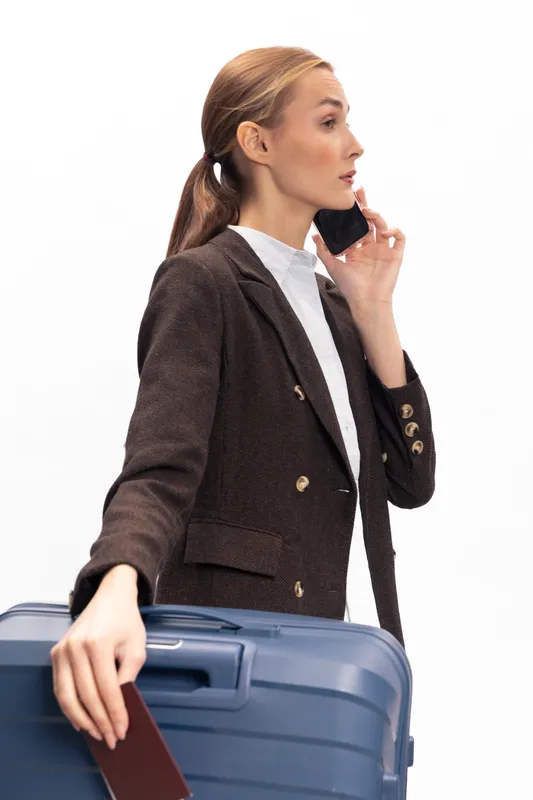 Young woman in brown coat holding passport and suitcase talking on phone. Concept of travel, business trip, and modern lifestyle with white background and studio lighting.