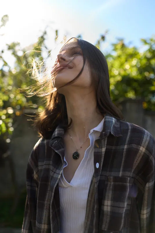 Woman lifestyle outdoors under sunlight, wind in hair, wearing plaid shirt and white top, relaxed pose, natural light, casual vibe on a bright day