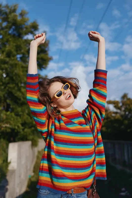 Woman in her 20s wearing a rainbow sweater and sunglasses, smiling with arms raised outdoors in a sunny suburban setting, casual outfit and joyful lifestyle fashion portrait.