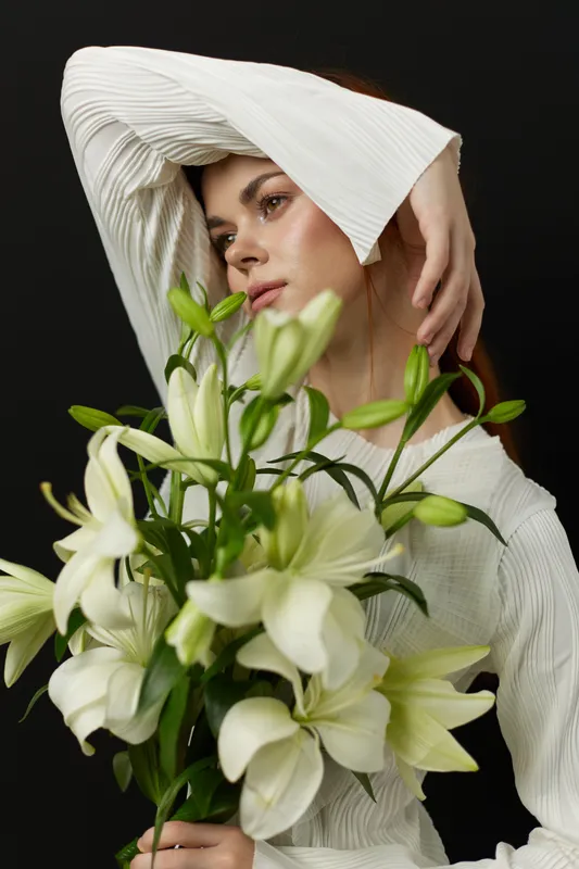 Elegant woman in a white blouse holding a bouquet of white lilies against a dark background, showcasing beauty and grace
