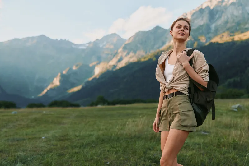 A young woman in casual clothes stands on a grassy field with mountains in the background, carrying a backpack. The scene represents outdoor lifestyle and adventure.