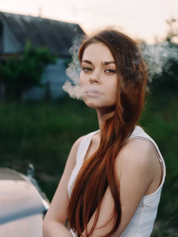 Young female model with long brown hair exhaling smoke, wearing a white tank top, with a natural outdoor background in soft light, evoking a sense of freedom and rebellion.