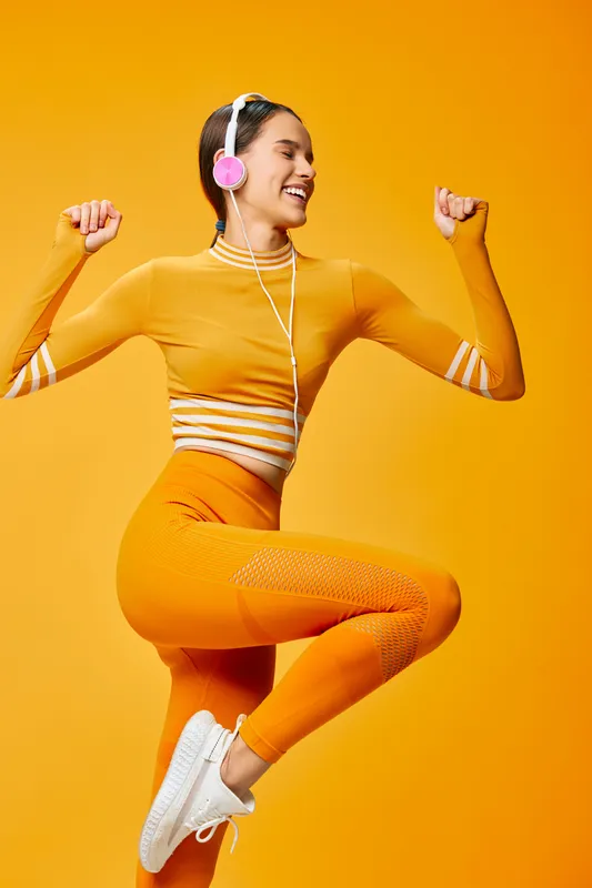 Vibrant fitness scene with a smiling woman in orange athletic wear jumping mid-air, wearing headphones; energetic workout moment captured in a bright studio backdrop.