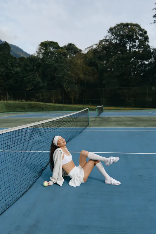 Young female tennis player resting on the court, wearing sporty attire, showcasing joy and confidence against a serene outdoor backdrop