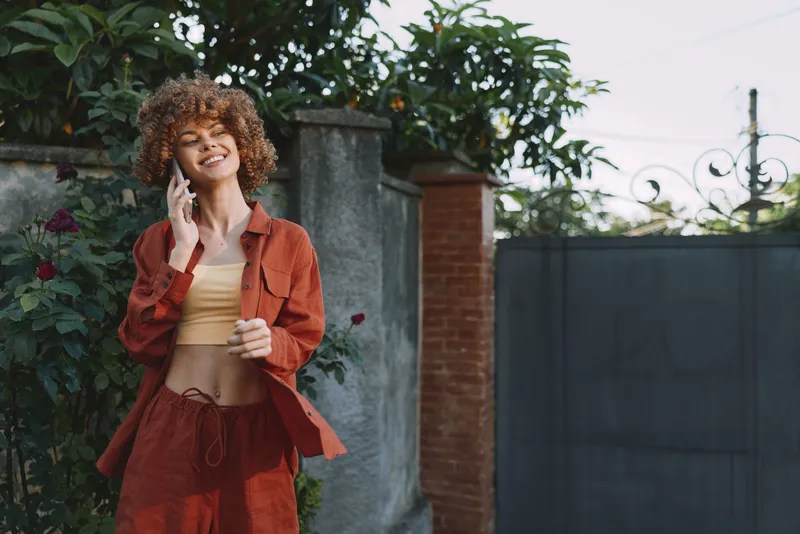 Smiling woman with curly hair wearing a rust colored jacket and matching outfit, talking on a phone outdoors in a sunny yard. Casual fashion, lifestyle mood, candid moment