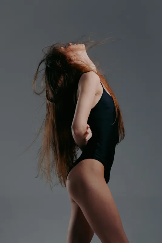 Woman with long red hair posing in black bodysuit against gray background, expressive pose, sensual mood, minimal studio setting.