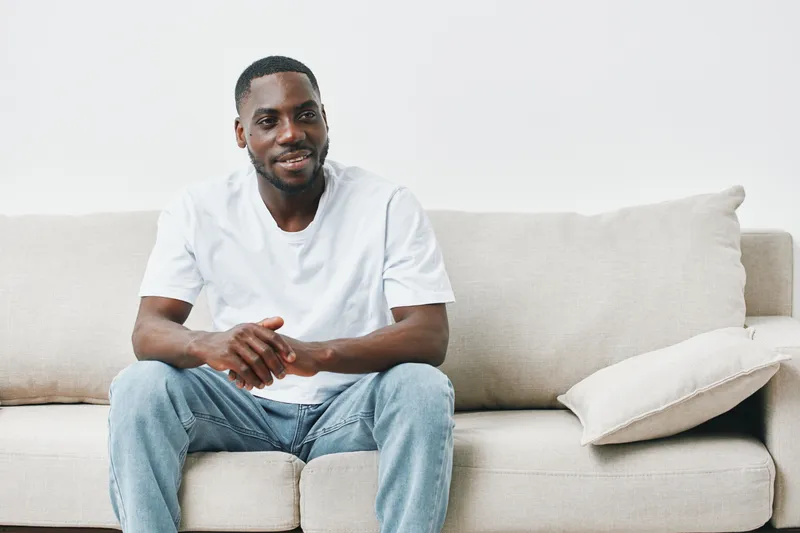 A smiling young Black man in a casual white t shirt and jeans sitting comfortably on a light sofa, radiating positivity and relaxation in a modern, minimalistic setting