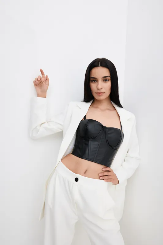 young woman in white suit and black corset posing confidently against white wall, fashion style with sleek straight hair and minimal makeup, modern look