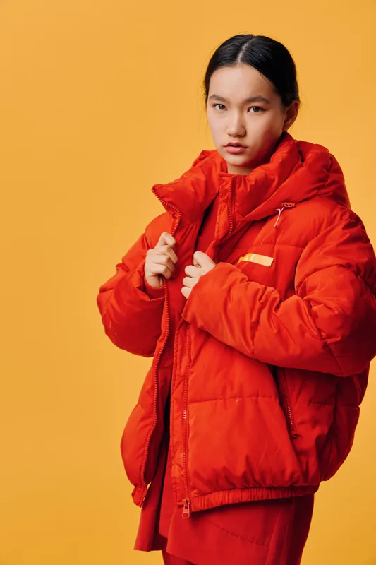 Young woman wearing bright red puffer jacket standing against yellow background, portrait with serious expression and casual fashion style for winter or autumn season.