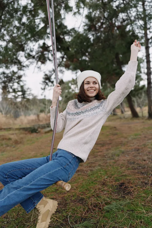 Woman on a swing outdoors, smiling and playful, wearing a knit sweater and beanie, surrounded by trees and grass in a calm countryside park under autumn light for a relaxed lifestyle moment.