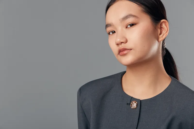 Young woman with ponytail wearing dark jacket looking at camera with neutral expression and natural makeup against gray studio background for portrait or fashion.