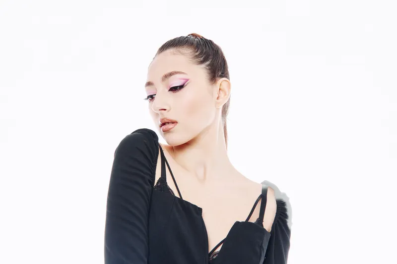 Young woman with bold pink eyeliner looking downward. Wearing black outfit with intricate straps, styled hair tied back. Creative beauty and fashion concept on white background.