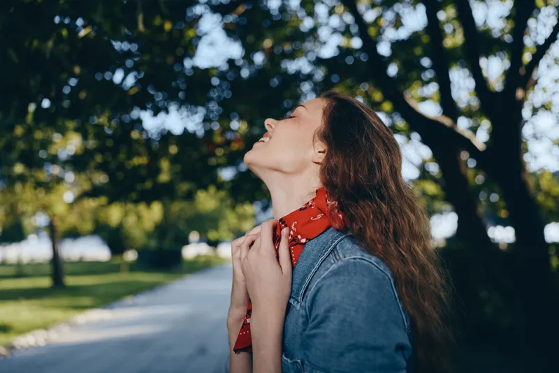 Young woman enjoying nature with closed eyes, wearing a denim jacket and red scarf, standing outdoors on a sunny day surrounded by green trees and pathway.