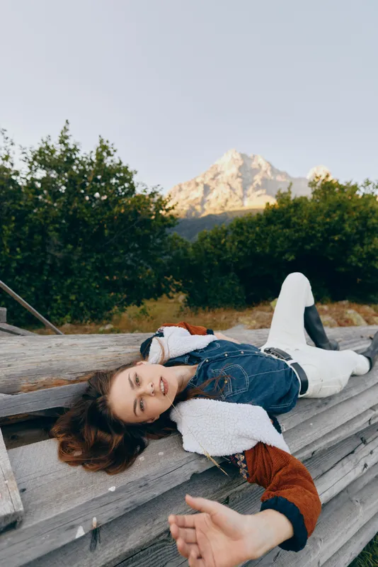 Woman lying on wooden logs outdoors near trees with mountain backdrop, portrait style, smiling and reaching hand toward the camera in a relaxed summer nature scene.