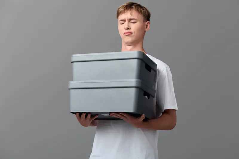Man carrying stackable storage boxes for moving, gray containers secured for relocation, clean studio setup, organized packing task, practical home or office transfer scenario