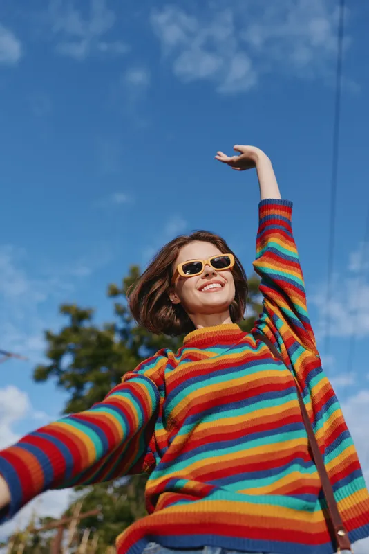 Woman in rainbow sweater wearing sunglasses smiling outdoors under blue sky, joyful portrait with raised arm, casual fashion and colorful striped knit in warm sunlight