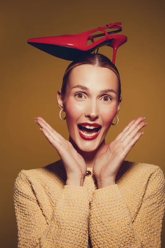 Happy woman with red high heel shoe on head smiles excitedly with hands near face. Studio portrait on brown background. Concept of fashion, fun, and playful mood.