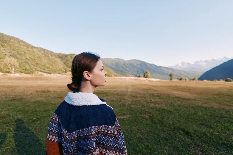 Woman in patterned sweater standing in meadow facing mountain landscape in profile. Outdoor nature scene with distant peaks, grassy field and clear sky at golden hour.