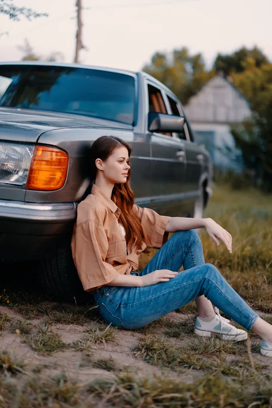 Young woman in casual outfit sits next to vintage car, enjoying a serene moment outdoors, embodying a relaxed summer vibe.