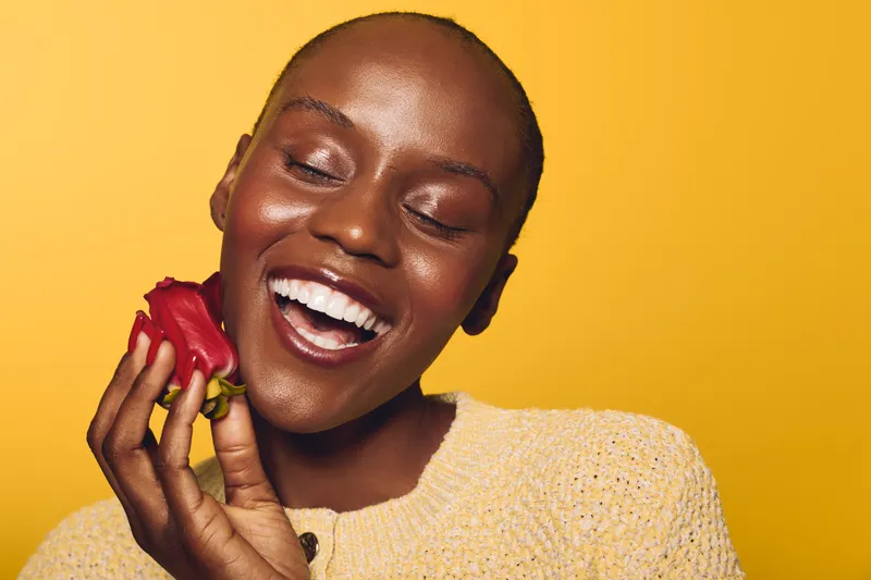 Happy dark skinned African woman with short hair holds red rose close to face, eyes closed and smiling. Yellow background and cozy beige sweater create warm joyful mood.