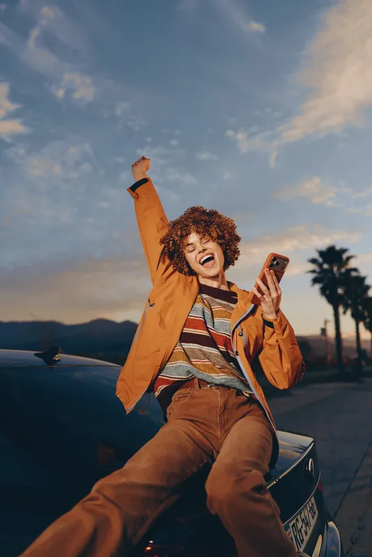 Woman lifestyle scene outdoors at sunset, joyful woman sitting on car hood holding smartphone, raising hand in motion, wearing warm jacket, distant palm trees, cinematic sky, roadtrip vibe