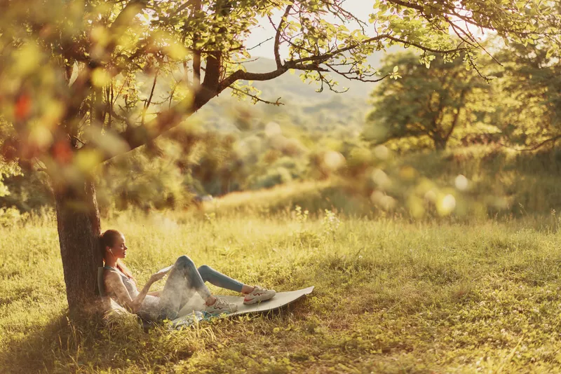 A young woman relaxing under a tree, enjoying a book in a serene green landscape during golden hour, capturing a sense of peace and connection with nature