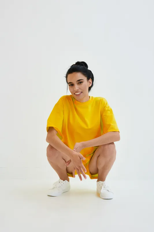 smiling woman in yellow oversized t-shirt and white sneakers crouching against white background, casual outfit, relaxed pose, youthful energy, studio lighting