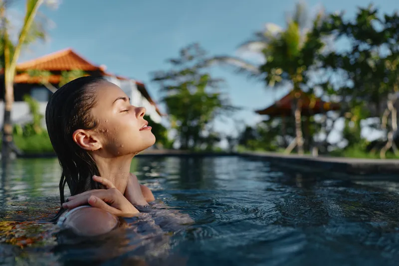 Young woman with wet hair relaxes peacefully in an outdoor pool surrounded by tropical greenery and traditional wooden pavilions under clear blue sky.