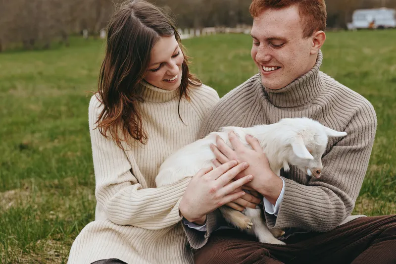 Couple enjoys a goats kid moment in a sunny meadow, sharing smiles and warmth, showcasing lifestyle and gentle affection as the white animal rests in their hands, creating a wholesome image of