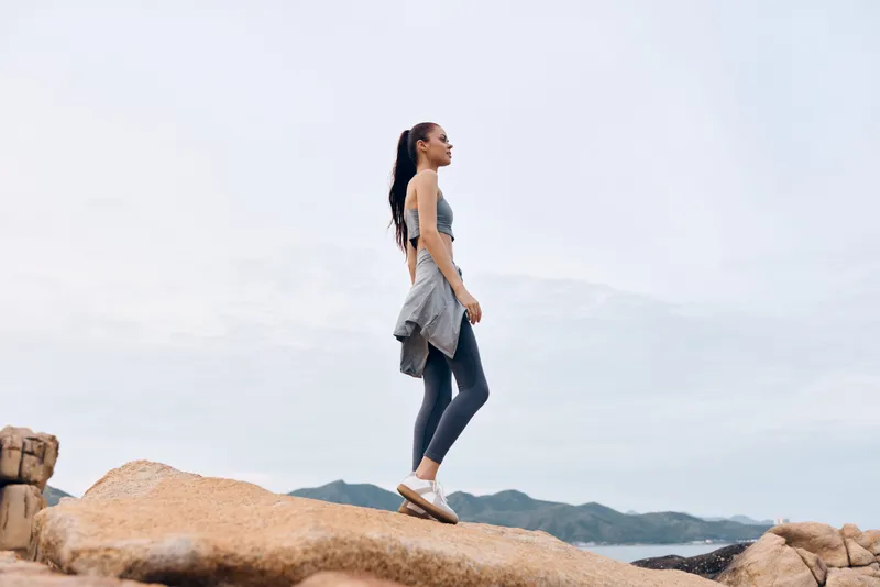 Young fit woman in sportswear stands confidently on rocky cliff, promoting healthy lifestyle, fitness, and outdoor activity with serene landscape background