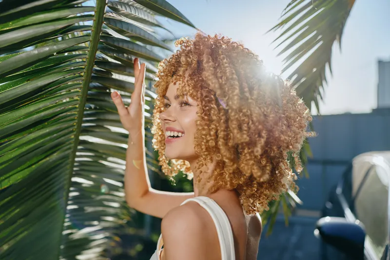 Smiling woman with curly hair enjoys sunny day near palm leaves, radiating happiness and natural beauty outdoors in warm light and vibrant summer atmosphere.