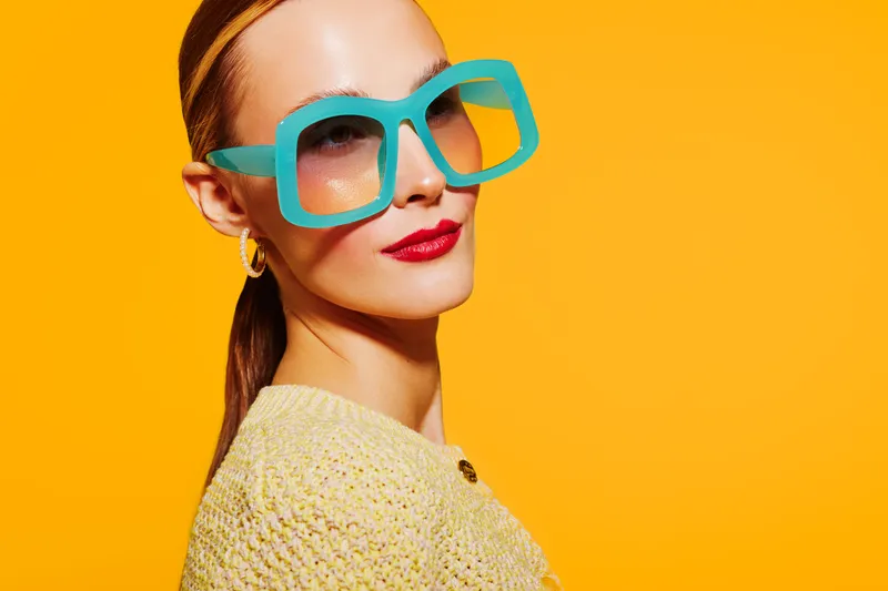 Stylish woman with large turquoise sunglasses and red lipstick poses confidently against bright yellow background. Fashion concept with bold colors and modern accessories.