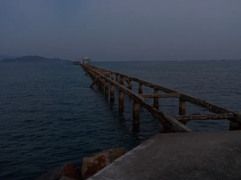 Old wooden pier extending into calm sea water during twilight, with a small shelter at the end and distant hills on the horizon, peaceful coastal scene