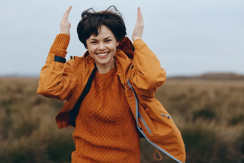 woman lifestyle outdoors, smiling in orange sweater, standing in autumn field, hands raised in wind, cheerful young adult, warm daylight, natural countryside background, relaxed pose