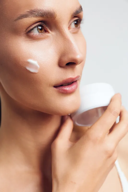 A woman applies facial cream to her cheek in soft, natural lighting. A skincare routine moment emphasizing hydration, self care, and calm beauty rituals.
