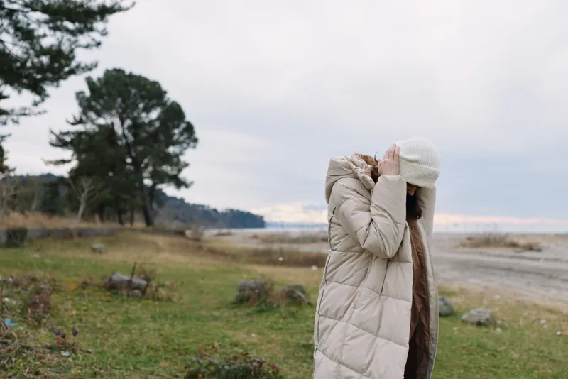 Woman in a light coat and white beanie stands outdoors near a coast, gazing at the horizon as wind blows, capturing a calm winter landscape and expansive seascape.