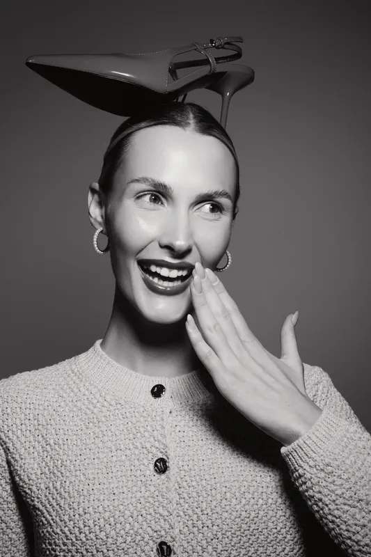 Smiling Caucasian woman with fair skin wears a high heel shoe on her head, covering her mouth with her hand. Concept of fashion, fun, and playful attitude in studio.