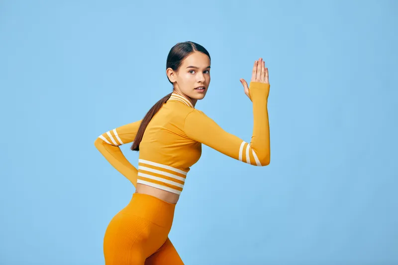 Sporty young woman in orange activewear posed in a dynamic running stance against a clean sky blue background, highlighting fitness, strength, athletic posture and expression.
