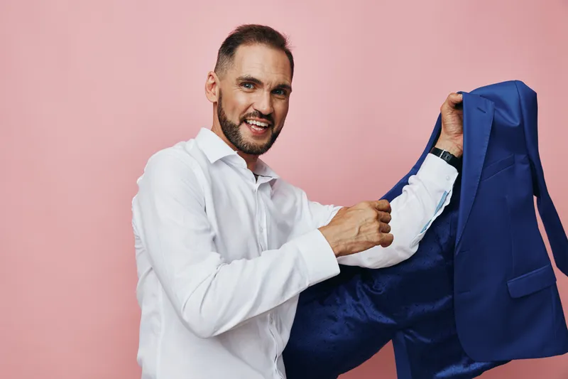 A professional businessman in a white shirt stands against a pink backdrop, holding a blue bag to convey confidence, readiness, and positive emotion in a clean, isolated color scene.