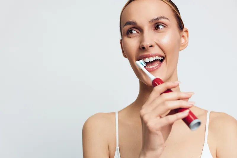 Young woman smiling while brushing teeth with electric toothbrush, clean white background, healthy dental care, morning routine, fresh and natural look