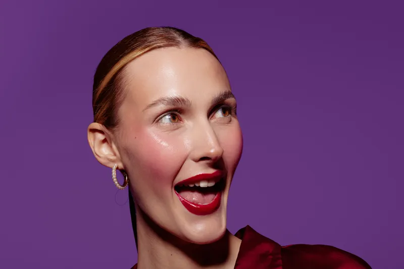 Happy woman with red lipstick and hoop earrings smiles joyfully on purple background. Concept of beauty, makeup, fashion, and positive emotions in studio portrait.