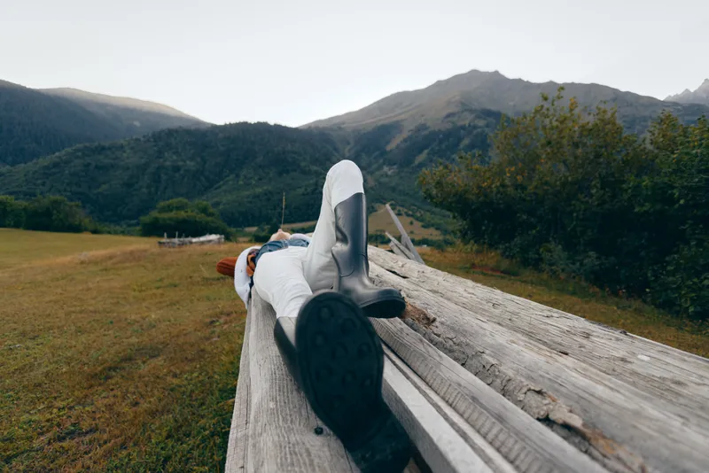 Person mountains meadow wooden relax outdoor lying on a rustic log bench in alpine field, traveler resting with boots and jacket, peaceful nature escape and wide scenic view.
