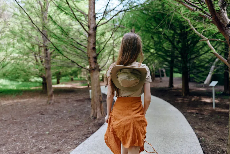 Woman walking on a park path with hat on her back, orange skirt and casual top, surrounded by trees and greenery. Outdoor stroll, summer fashion and peaceful nature escape.