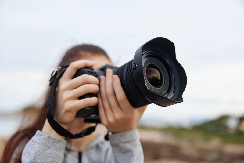 Young woman capturing a beautiful landscape with a DSLR camera, expressing joy and passion for photography outdoors, against a natural scenic background